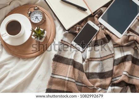 A cup of coffee, with an alarm clock tablet, a phone on a brown plaid on the bed. Top view with empty space for inscription