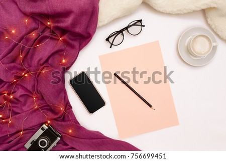 A cup of coffee, black smart, pink paper with new years goals. Scarf and Christmas lights on white background.