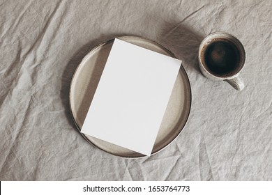 Cup Of Coffee And Blank Greeting Card, Invitation On Ceramic Plate. Moody Breakfast Table Mockup Scene. Beige Linen Tablecloth Background. Sparse Still Life Composition  Rustic Flat Lay, Top View.