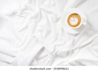 Cup Of Coffee Cappuccino In Bed On A White Sheet Of Bedding Linen. Cozy Soft Flatlay Morning Routine. Copy Space Mockup. Concept Minimalism.