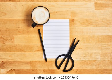 Cup of coffee or cappuccino with an empty sheet of paper, scissors and pencil on yellow wooden table. Top view. Copy space for text.
