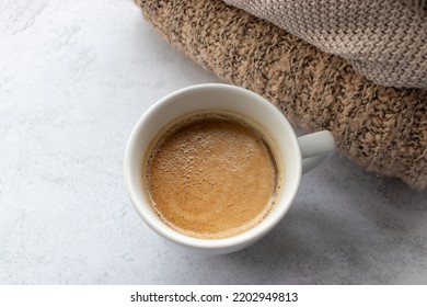 Cup Of Coffee Cappuccino, And Pile Of Beige Sweaters On White Table Background. Cozy Beige Autumn, Fall Aesthetic. Top View, Flat Lay, Copy Space

