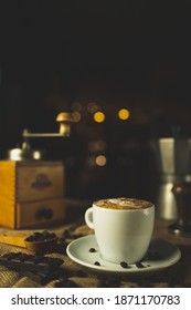 A Cup Of Coffee, Capuccino. A Nice Composition With A Coffee Grinder, An Espresso Maker And Some Chocolate. Restaurant And Coffee Shop. Nice Creamy Bokeh.