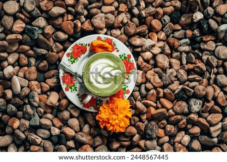 A cup of coffee with a floral design sits on a bed of rocks. There is a spoon on a saucer next to the cup. The coffee is green colored.