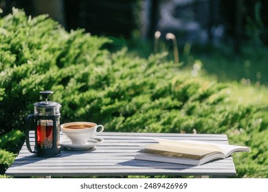 Cup of coffee, french press and an open book on a wooden table in a garden. Cozy picnic on backyard. Concepts of recreation area in public park or summer terrace in cafe, copy space backgrounds - Powered by Shutterstock