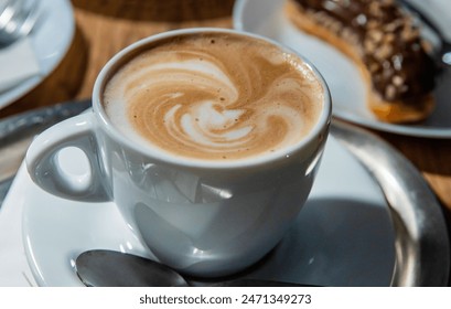 Cup of coffee latte with cream foam served on the white plate with spoon. Few plates on a wooden table in coffee shop. Mug of milk coffee, cappuccino, eclair with chocolate glaze. Selective focus - Powered by Shutterstock