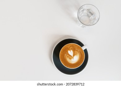 Cup Of Coffee On Black Saucer And Glass Of Water On White Background. Fresh Cappuccino With Beautiful Latte Art In Shape Of Heart. Coffee Break Concept. Top View, Flat Lay, Copy Space.