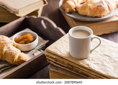 A cup of coffee on a stack of old books and a tray with croissants and jam on a wooden table. Relaxation, wellness and home reading.