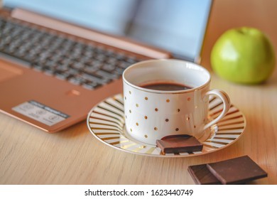 Cup of coffee on wood table with laptop on the background. Piece of black chocolate is on the saucer, coffee pause in the office for refreshment.