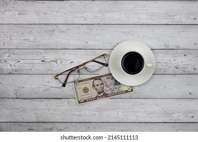 A Cup Of Coffee On A Wooden Table, A Five-dollar Bill, And Reading Glasses. Top View. The Concept Of Payment For Coffee.