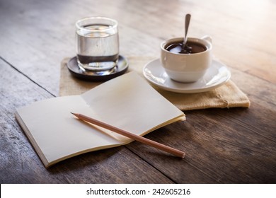 Cup Of Coffee On A Wooden Table With Glass Of Water On The Sackcloth Bags And Notebook And Pencil