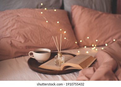 Cup of coffee with open paper book on wooden tray with liquid scented home fragrance in bed over glowing lights close up. Cozy atmosphere concept. Good morning.  - Shutterstock ID 1910755432