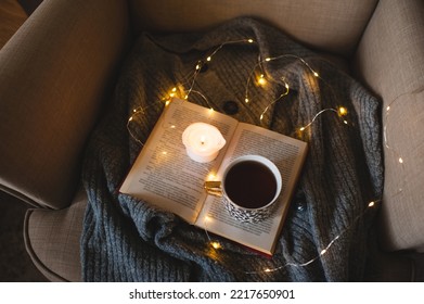 Cup Of Coffee With Paper Open Book And Scented Candle In Home Chair Over Christmas Lights Top View. 