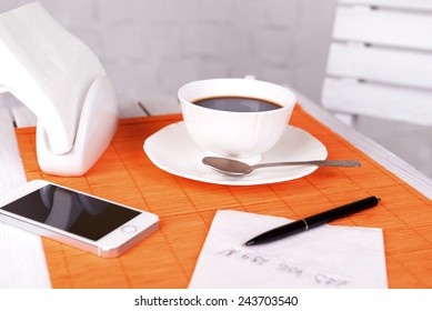 Cup Of Coffee With Pen And Phone Number On Napkin On Table With Orange Bamboo Mat, On White Wall Background