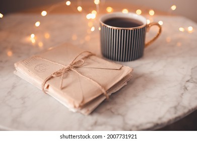 Cup Of Coffee With Pile Of Paper Handwritten Letters Stay On Marble Table In Room Over Glow Lights Close Up. Romantic Snail Mail Concept. Valentines Day. Cozy Atmosphere.