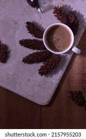 Cup of coffee and roasted coffee beans on a marble table surrounded by pine cones. Taken from a top view with a flat layout and dramatic lighting that casts elegant shadows.