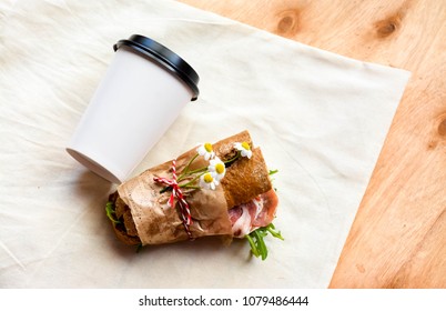 A Cup Of Coffee And A Sandwich Wrapped In Craft Paper On Rustic Wooden Table