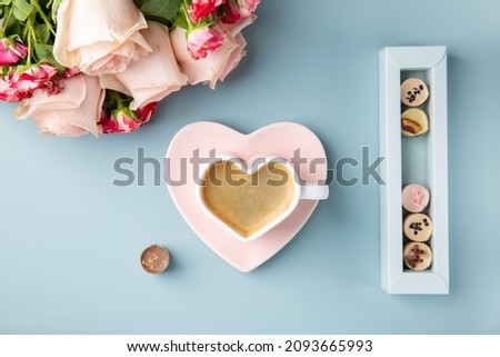Cup of coffee in shape of heart with chocolate candies and bouquet of roses on blue background. Love concept.  Top view.