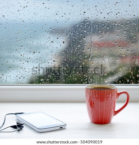 Cup of coffee, smartphone and headphones on a windowsill. In the background window with raindrops and clouds