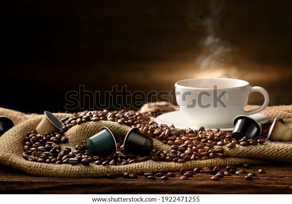 Cup of coffee with smoke\
and coffee beans and coffee capsules on burlap sack on old wooden\
background
