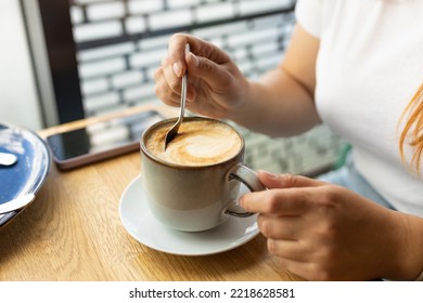 Cup Of Coffee With Spoon And Hand On Cafe Background. Young Woman Drinking Coffee Sitting At Wooden Table In Cafe In Daytime. Female's Hands Stir With Spoon Cappuccino Foam, Close-up.