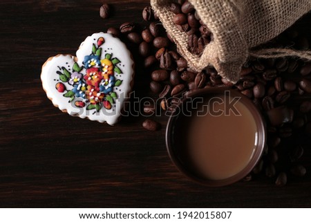 A cup of coffee is standing on a wooden table. Next to it lies a jute sack full of coffee and a decorated gingerbread heart.
