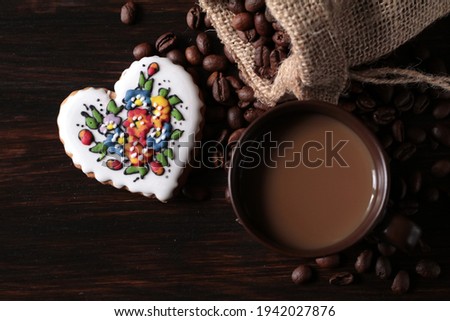 A cup of coffee is standing on a wooden table. Next to it lies a jute sack full of coffee and a decorated gingerbread heart.