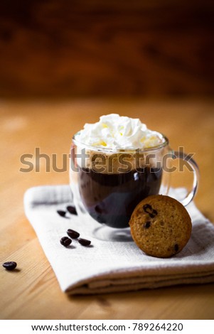 Cup of coffee with whipped cream topping on beige napkin in double botton glass with butter cookie on wooden brown table background