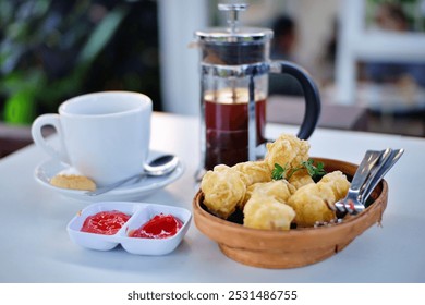 A cup of coffee in a white porcelain cup, a french press filled with coffee, a small bamboo basket with deep-fried tofu, and a small bowl of chili sauce on a white table in a cafe.  - Powered by Shutterstock