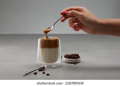 Cup with Dalgona coffee. Iced fluffy creamy whipped trend drink with coffee foam and milk in a glass with double glass. Female hand takes foam from a cup of coffee with a spoon. Shallow depth of field - Shutterstock ID 2187890539