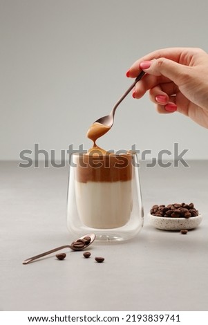 Cup with Dalgona coffee with spoon in a female hand. Iced fluffy creamy whipped trend drink with coffee foam and milk in a glass with double glass. Trendy Korean whipped coffee. Shallow depth of field