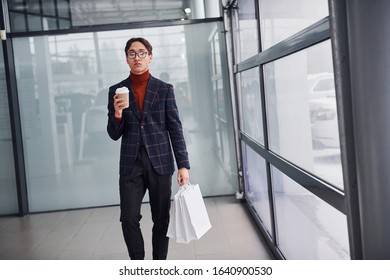 With cup of drink and shopping bags. Young business man in luxury suit and formal clothes is indoors in the office. - Shutterstock ID 1640900530