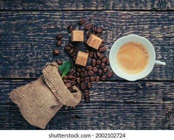 A Cup Of Frothy Coffee, A Bag Of Spilled Coffee Beans And Cane Sugar Lumps On An Old Wooden Surface. Flat Lay, Vintage