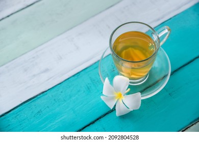 Cup of green tea with ginger servered with plumeria frangipani flower front of at sunset light on wooden tabletop. Sea view.