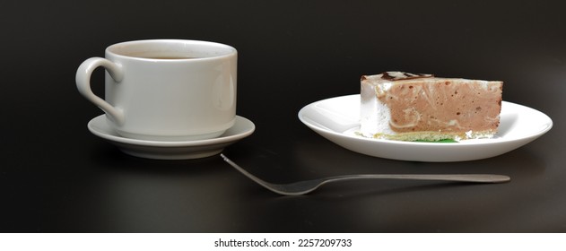 A cup of hot black coffee on a saucer, a fork and a plate with a piece of fresh cheesecake on a black background. Close-up.