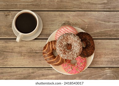 A cup of hot black coffee on a saucer and a plate with a bunch of fresh donuts in multi-colored glaze on a wooden table. Top view, flat lay. - Powered by Shutterstock