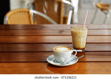 A cup of hot cappuccino and a glass of cold iced latte on wooden table in a street coffee shop against blurred background.