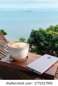A cup of hot cappuchino and cell phone on wooden brown board over seascape background. Fresh coffee and good morning concept.