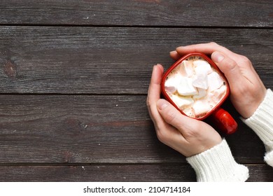 Cup hot chocolate with marshmallows in the shape of hearts and woman hands in white sweater on brown wooden background. Valentine's Day celebration. Top view with copy space