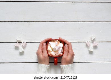 Cup hot chocolate with marshmallows in the shape of hearts and woman hands in white sweater on white wooden background. Valentine's Day celebration. Top view with copy space