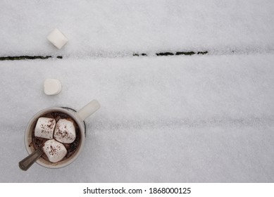 Cup Of Hot Cocoa With Marshmallows On A Snow Covered Picnic Table.