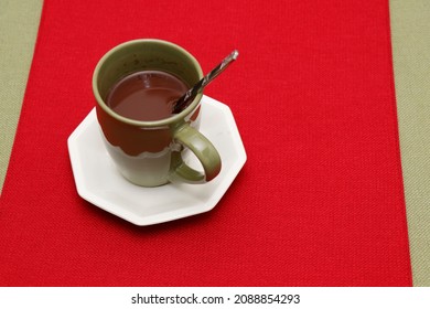 A cup of hot cocoa mix with spoon on a white saucer placed on a red place mat on a green tablecloth. Warm hot chocolate mix in a cup with a metal spoon on a red and green textile tabletop background.