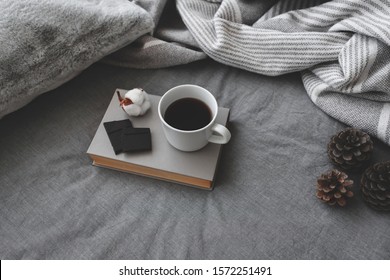 Cup Of Hot Coffee, Dark Chocolate, Book, Blanket, Pillow, Plaid, Cotton Flower And Pine Cones Lie On The Bed With Gray Linens. Still Life Details. Winter Scene. Cozy Concept. Dark Grey Colors.