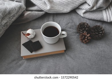 Cup Of Hot Coffee, Dark Chocolate, Book, Blanket, Pillow, Plaid, Cotton Flower And Pine Cones Lie On The Bed With Gray Linens. Still Life Details. Winter Scene. Cozy Concept. Dark Grey Colors.