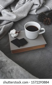 Cup Of Hot Coffee, Dark Chocolate, Book, Blanket, Plaid, Cotton Flower And Pine Cones Lie On The Bed With Gray Linens. Still Life Details. Winter Scene. Cozy Concept. Dark Grey Colors.