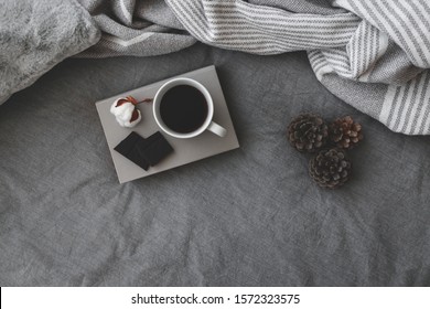 Cup Of Hot Coffee, Dark Chocolate, Book, Blanket, Plaid, Cotton Flower And Pine Cones Lie On The Bed With Gray Linens. Winter Scene. Cozy Concept. Dark Grey Colors. Flat Lay, Top View. Copy Space.