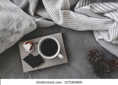 Cup Of Hot Coffee, Dark Chocolate, Book, Blanket, Fluffy Pillow, Plaid, Cotton Flower And Pine Cones Lie On The Bed With Gray Linens. Winter Scene. Cozy Concept. Dark Grey Colors. Flat Lay, Top View.