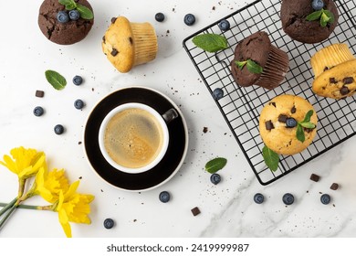 Cup of hot coffee, fresh baked muffins with chocolate chips, blueberry berries and mint leaves on white marble table background. Chocolate  cupcake closeup as dessert for coffee break. Top view.