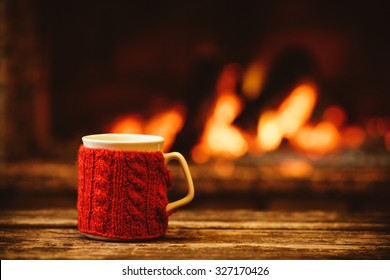 Cup Of Hot Drink In Front Of Warm Fireplace. Holiday Christmas Concept. Mug In Red Knitted Mitten Standing Near Fireside. Cozy Relaxed Magical Atmosphere In A Chalet.