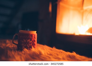 Cup Of Hot Drink In Front Of Warm Fireplace. Holiday Christmas Concept. Mug In Red Knitted Mitten Standing Near Fireside. Cozy Relaxed Magical Atmosphere In A Chalet.